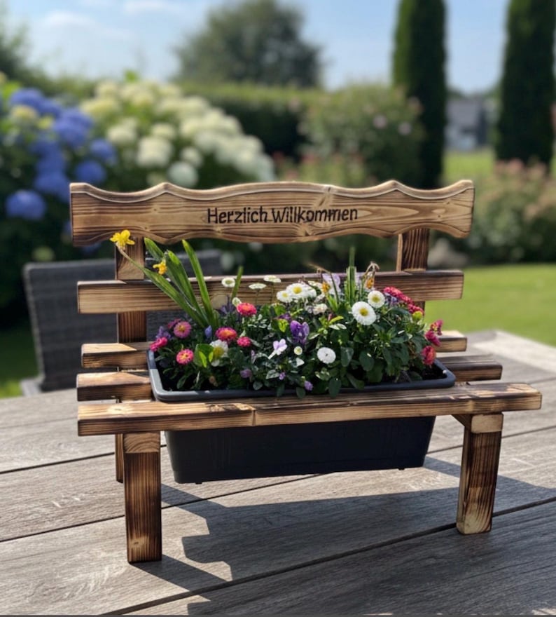 Puede incluir: Una jardinera en forma de banco de madera con un contenedor de pl&aacute;stico negro lleno de flores coloridas. El banco tiene un respaldo con la inscripci&oacute;n "Herzlich Willkommen" y est&aacute; sobre una superficie de madera.