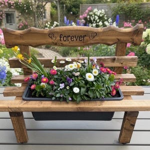 Puede incluir: Un banco de madera con una jardinera llena de flores coloridas. El banco tiene un respaldo con la palabra "forever" grabada. Las flores incluyen margaritas y pensamientos. El banco est&aacute; al aire libre.