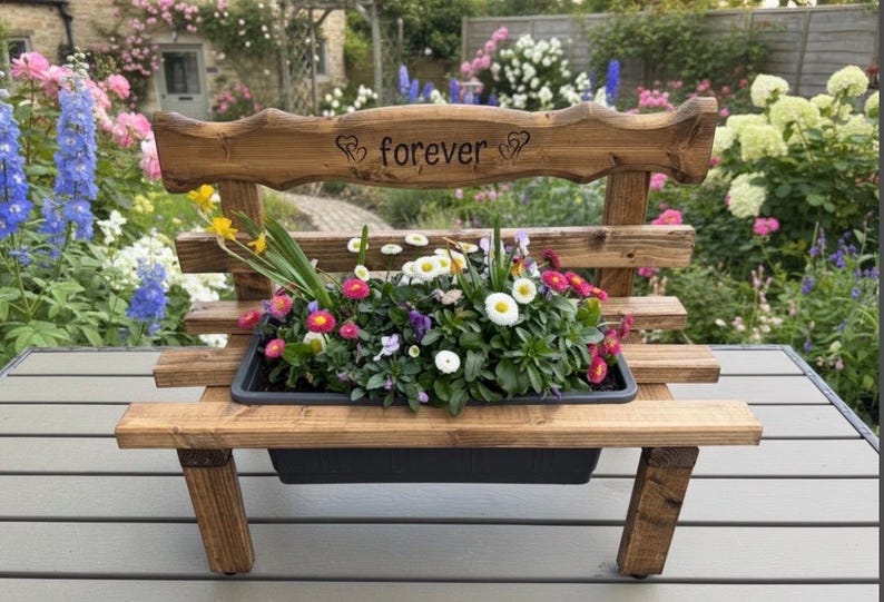 Puede incluir: Un banco de madera con una jardinera llena de flores coloridas. El banco tiene un respaldo con la palabra "forever" grabada. Las flores incluyen margaritas y pensamientos. El banco est&aacute; al aire libre.