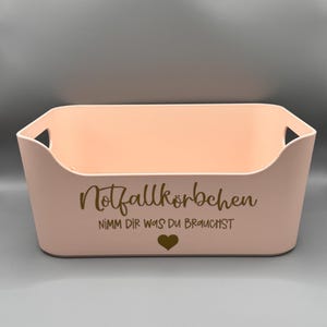 Könnte beinhalten: Ein rosa Plastik-Aufbewahrungskorb mit dem Text "Notfallkörbchen NIMM DIR WAS DU BRAUCHST" und einem kleinen Herz in goldener Schrift.