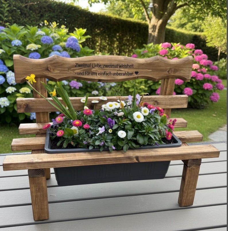 Puede incluir: Banco de jard&iacute;n de madera con jardinera incorporada llena de flores variadas. El banco est&aacute; te&ntilde;ido de marr&oacute;n oscuro y tiene un respaldo decorativo con texto grabado. Las flores incluyen margaritas y otras flores coloridas.
