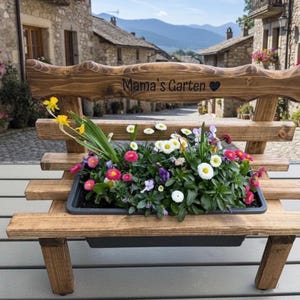 Puede incluir: Un banco de madera con una jardinera llena de flores coloridas. El banco tiene un respaldo con las palabras "Mama's Garten" y un coraz&oacute;n tallado. El banco es de madera marr&oacute;n claro y est&aacute; al aire libre.