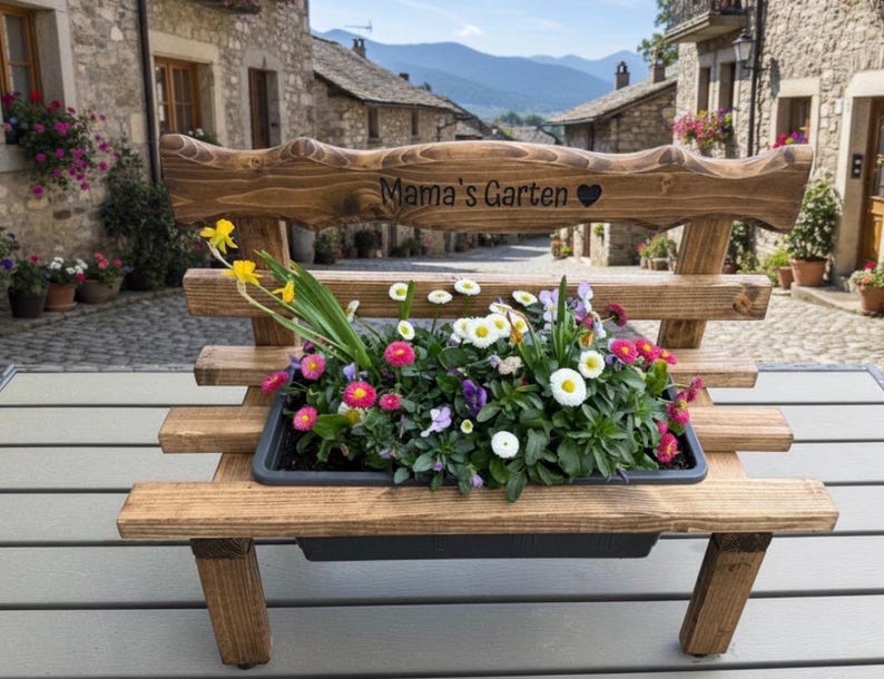 Puede incluir: Un banco de madera con una jardinera llena de flores coloridas. El banco tiene un respaldo con las palabras "Mama's Garten" y un coraz&oacute;n tallado. El banco es de madera marr&oacute;n claro y est&aacute; al aire libre.