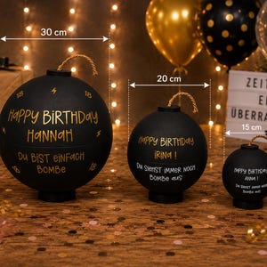 Könnte beinhalten: Drei kugelförmige, schwarze Behälter in Bombenform mit goldener Schrift und einer Kordel-Lunte oben. Der größte misst 30 cm, der mittlere 20 cm und der kleinste 15 cm. Jeder Behälter trägt personalisierte Geburtstagsbotschaften auf Deutsch mit Namen.