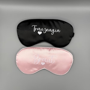 Könnte beinhalten: Zwei Satin-Schlafmasken, eine schwarze und eine rosa. Die schwarze Maske hat den Text "Trauzeugin" mit einem weißen Herzen. Die rosa Maske hat den Text "Braut" mit einem weißen Herzen.