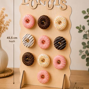 Könnte beinhalten: Ein Donut-Ständer aus hellem Holz mit dem Schriftzug DONUTS an der Oberseite. Er verfügt über zehn Stifte, an denen verschiedene glasierte Donuts hängen. Der Ständer ist 45,5 cm hoch, 26,5 cm breit und 12 cm tief und steht auf zwei Stützfüßen.