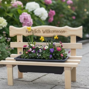 Könnte beinhalten: Eine kleine Holzbank mit einer schwarzen Pflanzschale, die mit bunten Blumen gefüllt ist. Die Bank hat eine Rückenlehne mit dem eingravierten Schriftzug "Herzlich Willkommen". Der Hintergrund zeigt verschiedene blühende Blumen.