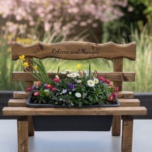 Puede incluir: Un peque&ntilde;o banco de madera oscura que sirve como jardinera, lleno de flores coloridas. El respaldo lleva la inscripci&oacute;n "Rebecca und Marwin". La jardinera negra contiene una variedad de flores vibrantes, ideal para un jard&iacute;n.