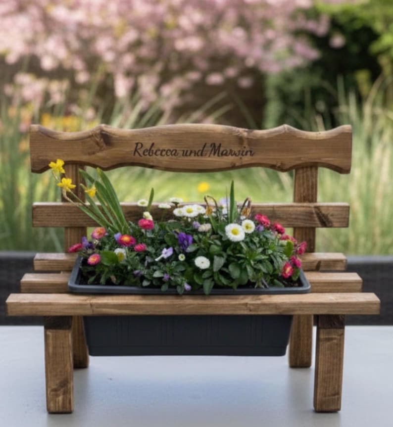 Puede incluir: Un peque&ntilde;o banco de madera oscura que sirve como jardinera, lleno de flores coloridas. El respaldo lleva la inscripci&oacute;n "Rebecca und Marwin". La jardinera negra contiene una variedad de flores vibrantes, ideal para un jard&iacute;n.