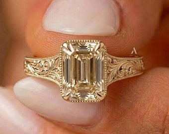 Pode incluir: Um anel de ouro com uma grande pedra preciosa cor de champanhe em corte esmeralda, engastada numa moldura. A banda é feita de ouro amarelo e apresenta padrões de volutas gravados intrincados nos ombros, estendendo-se a partir da pedra central.