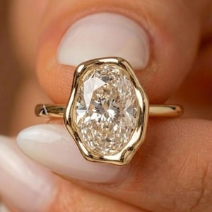 May include: A gold ring featuring a large, oval-cut, clear gemstone set in a unique, organic-shaped, polished gold bezel setting. The band is thin and smooth, crafted from yellow gold, highlighting the contrast between the faceted stone and the metal.