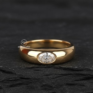 Puede incluir: Un anillo de oro con un acabado pulido, con un diamante de talla ovalada engastado en el centro. El anillo se muestra sobre una superficie oscura y texturizada, resaltando el diseño y el brillo del anillo.