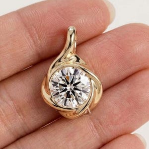 May include: A gold pendant featuring a single round brilliant-cut clear gemstone set in a swirling, polished metal frame. The design includes a tapered bail at the top, creating a continuous, fluid shape that highlights the faceted stone.