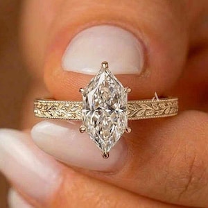 Puede incluir: Un anillo de compromiso de oro con un gran diamante talla marquesa. La banda presenta un intrincado patrón floral grabado. El anillo se sostiene, mostrando el brillo del diamante y el diseño detallado de la banda.