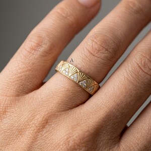 May include: A gold band ring featuring a repeating fan-shaped pattern engraved across the surface. Each fan motif contains small, clear, sparkling stones set within the triangular sections, creating a textured and detailed metallic jewelry piece.