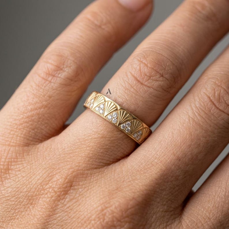 May include: A gold band ring featuring a repeating fan-shaped pattern engraved across the surface. Each fan motif contains small, clear, sparkling stones set within the triangular sections, creating a textured and detailed metallic jewelry piece.