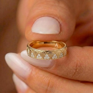 May include: A gold band ring featuring a repeating fan-shaped pattern engraved along the exterior. Each fan motif is accented with small, clear, round-cut stones embedded within the design. The metal has a polished, reflective yellow gold finish.