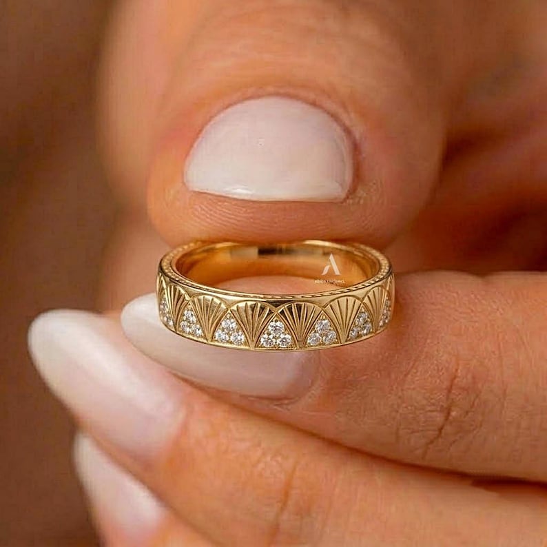 May include: A gold band ring featuring a repeating fan-shaped pattern engraved along the exterior. Each fan motif is accented with small, clear, round-cut stones embedded within the design. The metal has a polished, reflective yellow gold finish.