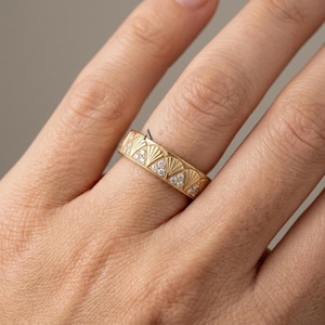 May include: A gold band ring featuring a repeating pattern of engraved fan-shaped motifs. Each fan design is accented with three small, clear, round-cut stones set within the lower triangular section of the pattern, creating a textured look.