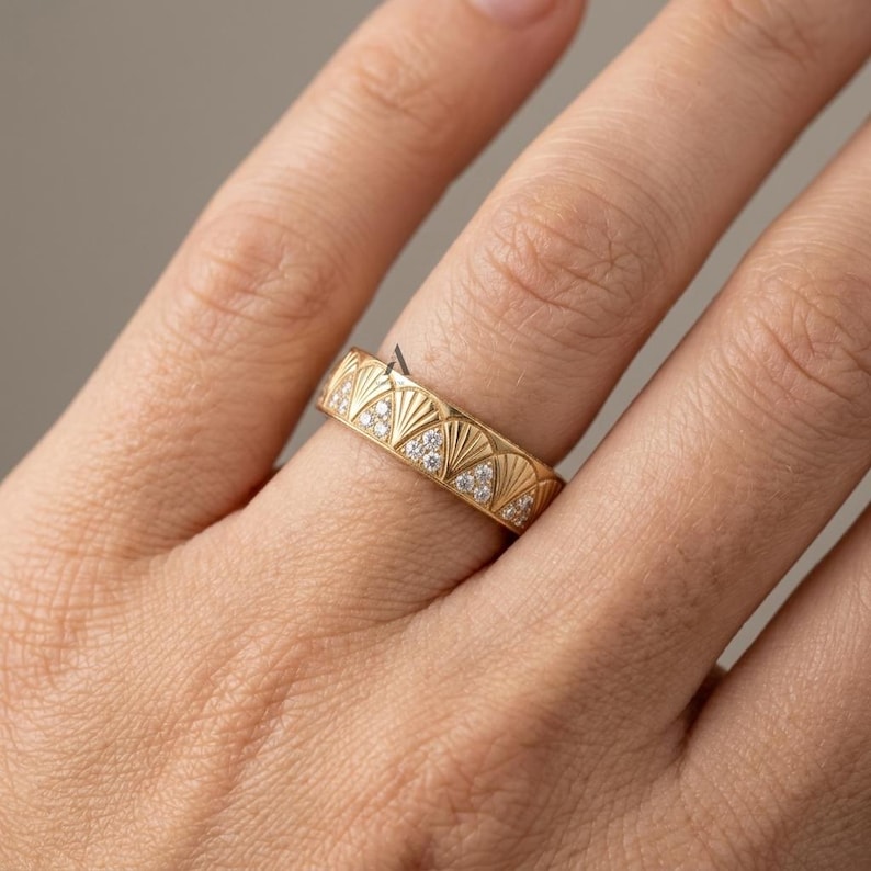 May include: A gold band ring featuring a repeating pattern of engraved fan-shaped motifs. Each fan design is accented with three small, clear, round-cut stones set within the lower triangular section of the pattern, creating a textured look.