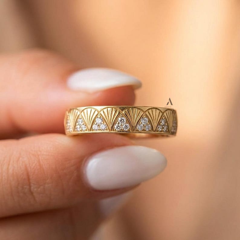 May include: A gold band ring featuring a repeating fan-shaped pattern engraved across the surface. Small, clear, round-cut stones are set within the base of each fan motif, creating a consistent, decorative design around the entire circumference.