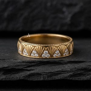 May include: A gold band ring featuring a repeating pattern of engraved fan-shaped motifs alternating with triangular sections inlaid with small, clear, round-cut stones. The metal surface has a polished finish with a consistent yellow gold tone.