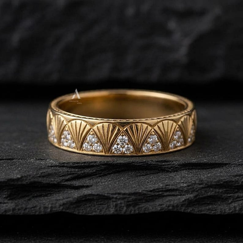 May include: A gold band ring featuring a repeating pattern of engraved fan-shaped motifs alternating with triangular sections inlaid with small, clear, round-cut stones. The metal surface has a polished finish with a consistent yellow gold tone.