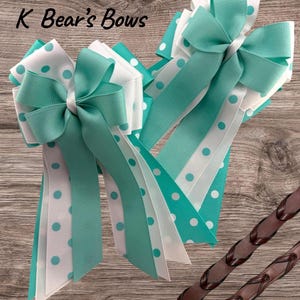 May include: Two large hair bows with turquoise, white, and polka dot ribbons. The bows have long, layered tails and are embellished with a bow knot. The text "K Bear's Bows" is visible in the upper left corner.