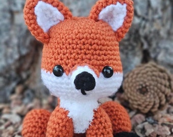 May include: A handmade crochet fox doll with orange, white, and black details. The fox has large, round black eyes, a white muzzle, and a black nose. The ears are orange with white inner ear details. The fox is sitting on a bed of small rocks.