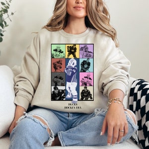 Op de afbeelding: Een beige sweatshirt met een afbeelding van een collage van hockey spelers in verschillende gekleurde vierkanten. De tekst "Ducks Hockey Era" is onder de collage gedrukt.