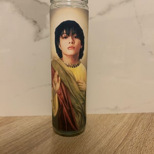 May include: A glass candle with a printed image of a person with black hair wearing a yellow robe with a red and green sash. The person is looking directly at the viewer.