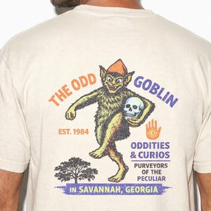 May include: A beige t-shirt featuring a graphic on the back of a goblin wearing an orange hat and holding a skull. The text reads THE ODD GOBLIN, EST. 1984, ODDITIES & CURIOS, PURVEYORS OF THE PECULIAR, IN SAVANNAH, GEORGIA, with a tree and hand icon.