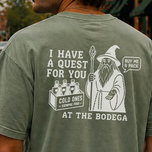May include: A sage green t-shirt featuring a white pixelated graphic on the back. The design shows a wizard holding a staff next to a crate of bottles labeled COLD ONES SIEMPRE FRIO. Text reads I HAVE A QUEST FOR YOU AT THE BODEGA and BUY ME 6 PACK.