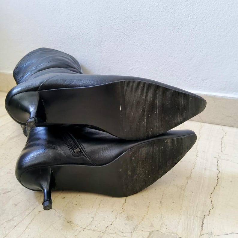 May include: Black leather knee-high boots with pointed toes and stiletto heels. The boots have a side zipper and a textured sole. The heels are approximately 2 inches (5 cm) high. The boots are lying on a light-colored surface.