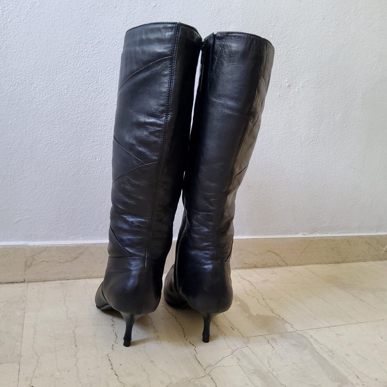 May include: A pair of black leather knee-high boots with a pointed toe and a stiletto heel. The boots have a zipper closure and a subtle stitched design on the front. The boots are made of a smooth, shiny leather.