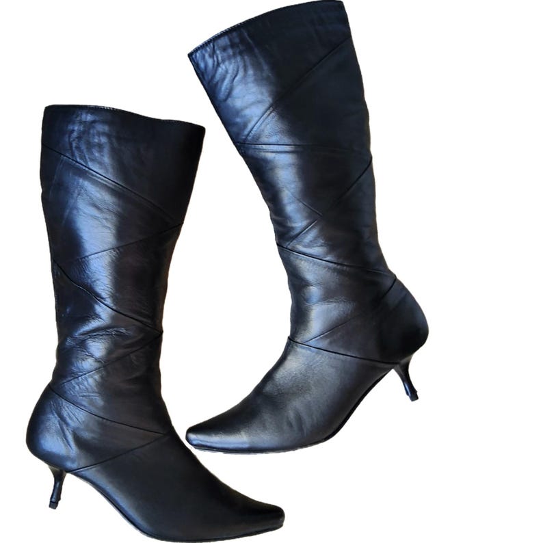 May include: A pair of black leather knee-high boots. The boots have a pointed toe and a small, curved heel. Diagonal stitching details run up the front of the boots. The boots are set against a white background.