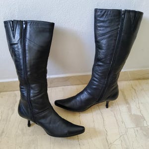 May include: A pair of black leather knee-high boots with pointed toes and small heels. Each boot has a zipper running along the side. The boots are made of a smooth, shiny leather.