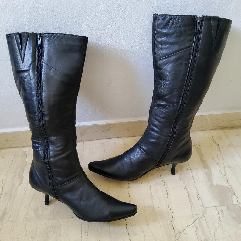 May include: A pair of black leather knee-high boots with pointed toes and small heels. Each boot has a zipper running along the side. The boots are made of a smooth, shiny leather.