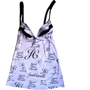 Puede incluir: Una camisola blanca con tirantes y ribetes negros, con el texto "Just Cavalli" y las iniciales "JC" en un patrón repetitivo. El escote tiene forma de V profunda.