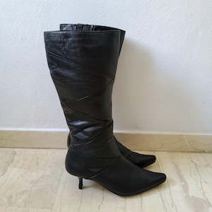 May include: A pair of black leather knee-high boots with a pointed toe and a small heel. The boots have a decorative stitched design on the front and a zipper closure. The boots are standing on a light-colored surface.