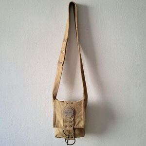 Op de afbeelding: Tan canvas crossbody tas met een verstelbare riem. De tas heeft een voorflap met een metalen gespsluiting en een cirkelvormige patch met de tekst "1978 DIESEL". De tas hangt tegen een witte muur.