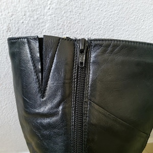 May include: Close-up of a black leather boot with a zipper closure. The boot features a V-shaped design at the top and visible stitching along the seams. The zipper is silver-toned and runs vertically down the side of the boot.