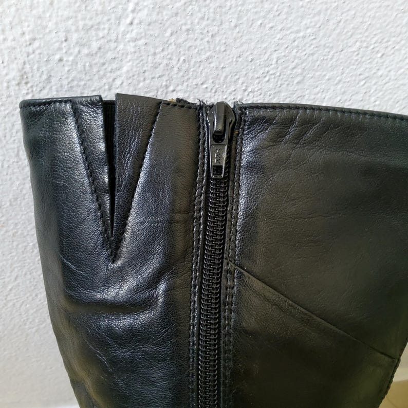 May include: Close-up of a black leather boot with a zipper closure. The boot features a V-shaped design at the top and visible stitching along the seams. The zipper is silver-toned and runs vertically down the side of the boot.