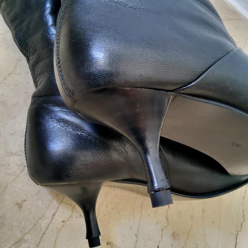 May include: Black leather boots with a pointed toe and a small, curved heel. The boots have a smooth, polished finish and a quilted design on the upper shaft. The heels are approximately 2 inches (5 cm) high.