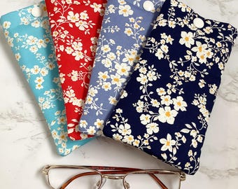 May include: Four fabric eyeglass cases in shades of blue, red, and navy, each decorated with a white floral pattern. A pair of gold-rimmed eyeglasses rests below the cases, providing a complete eyewear storage solution.