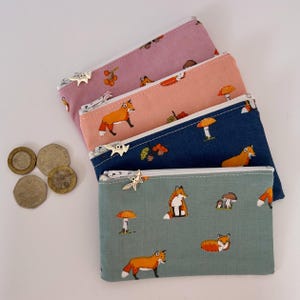 May include: Four rectangular coin purses in pink, peach, navy, and green, each with a white zip and a star-shaped pull. The purses feature a repeating pattern of foxes and mushrooms. Several coins are visible to the left of the purses.