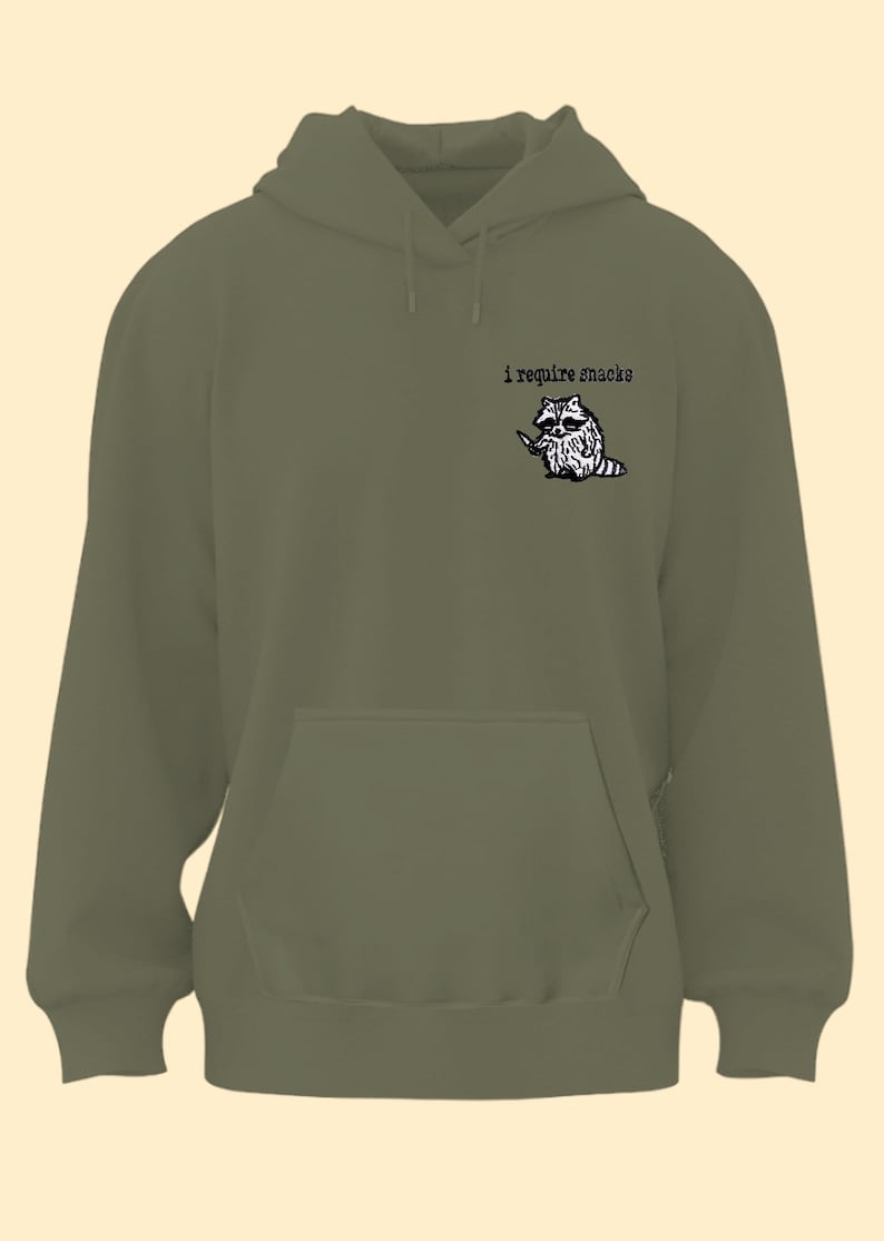 May include: Olive green hoodie with a kangaroo pocket and drawstring hood. The upper left chest features a black and white illustration of a raccoon holding a knife, with the text "i require snacks" above it.