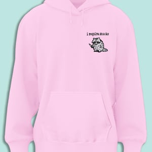 May include: A light pink hoodie with a kangaroo pocket and drawstring hood. The upper left chest features the text "i require snacks" above a black and white illustration of a raccoon holding a knife.