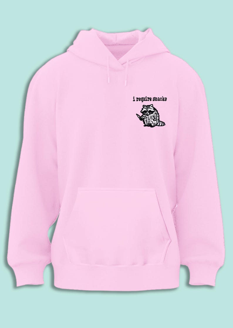 May include: A light pink hoodie with a kangaroo pocket and drawstring hood. The upper left chest features the text "i require snacks" above a black and white illustration of a raccoon holding a knife.