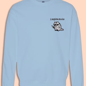 May include: Light blue sweatshirt featuring a graphic of a raccoon holding a knife, with the text "i require snacks" above the raccoon. The raccoon is black and white with a pink and white striped tail. The sweatshirt is a crew neck.
